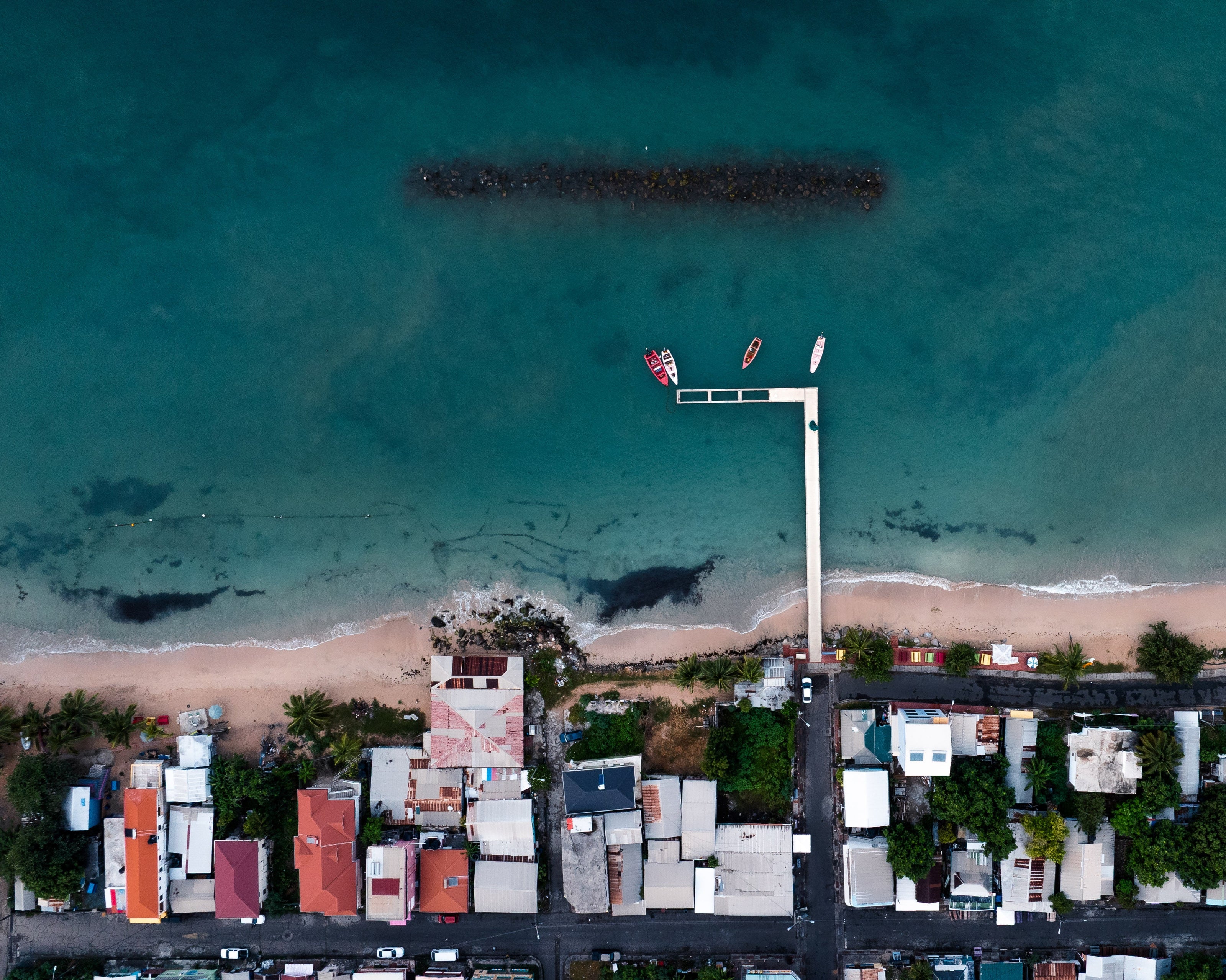 A photograph taken from a DJI Mavic 3 Pro drone overlooking the St Lucian coastline. The image centers around a small dock with a break built to protect fishing boats from incoming waves.
